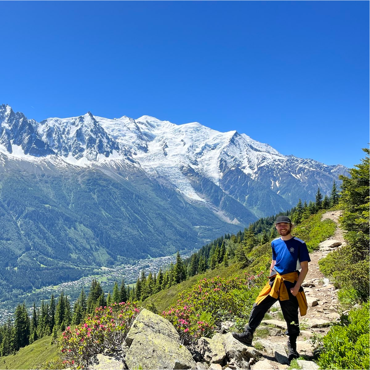 Me posing in front of a forest, valley, and mountain range while hiking in Switzerland on a beautiful sunny day